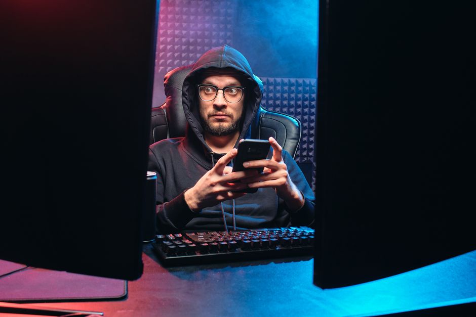 A man in a hoodie using a smartphone, surrounded by tech gear in a dimly lit room.