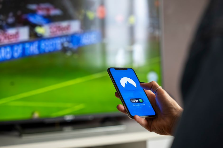 Close-up of a person holding a smartphone with a VPN app, streaming sports on TV.