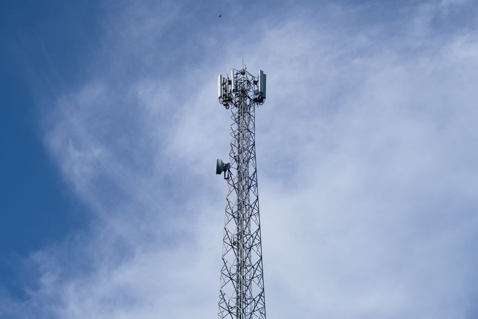 A tall metal communications tower stands against a backdrop of a clear blue sky with scattered clouds.