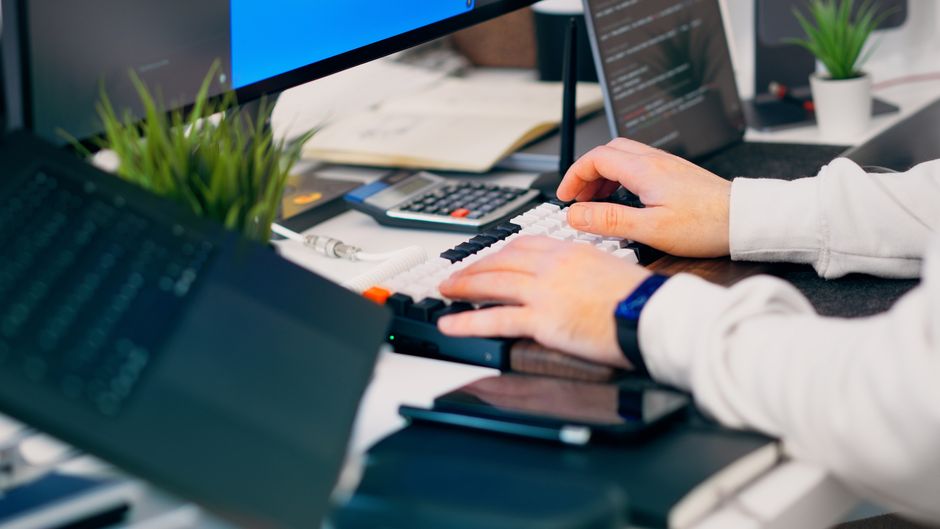 A close-up of a person typing on a keyboard in a modern tech workspace with gadgets and a monitor.